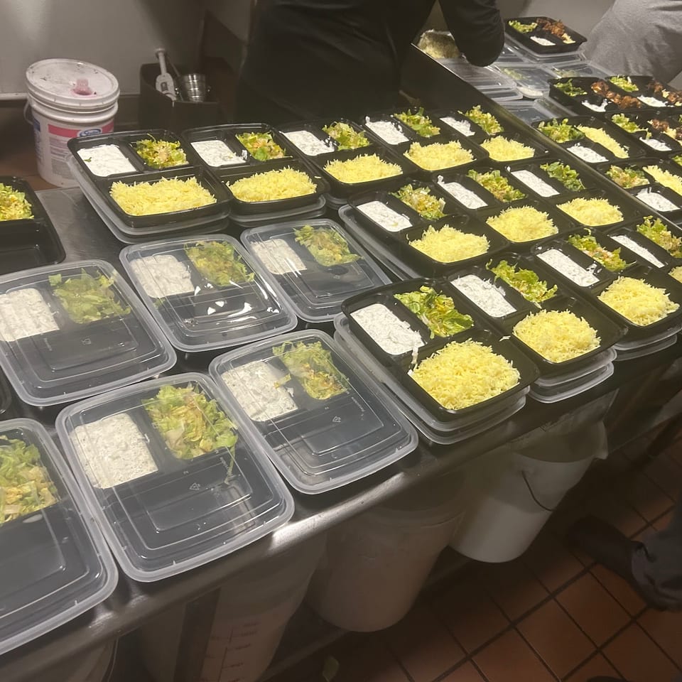 Numerous meal prep containers filled with rice, salad, and sauce, some with lids, on a metal counter.
