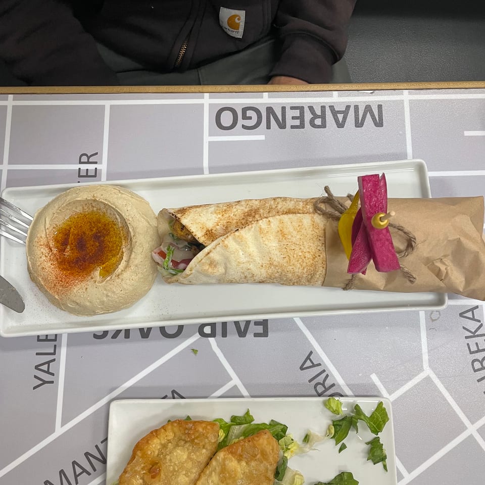 Hummus with paprika and olive oil, a wrapped falafel, and two fried pastries on a map-patterned table.