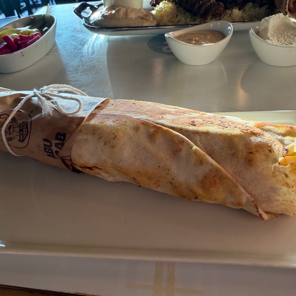 A large Abu Kebab wrap, tied with twine, on a white plate with pickles and dips.