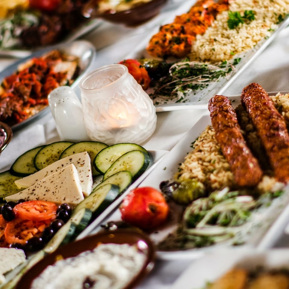 A table laden with various Mediterranean dishes, including kebabs, rice, dips, cheese, and vegetables.