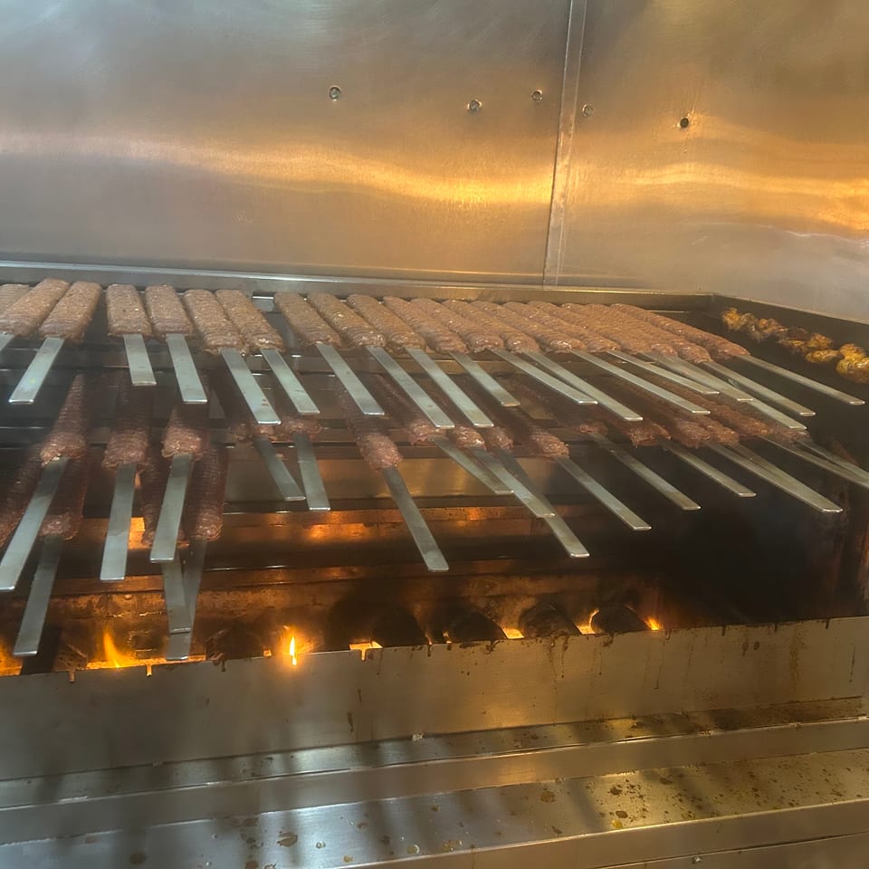 Numerous skewers of meat cooking over an open flame grill in a commercial kitchen.