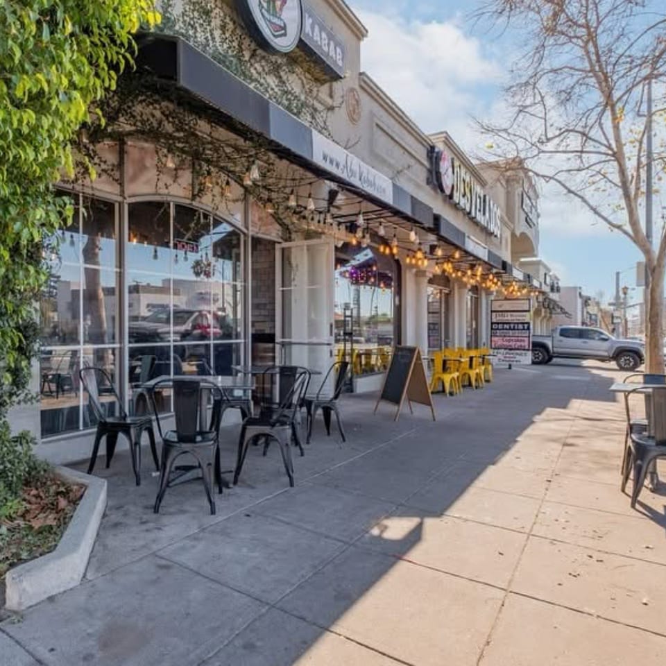 Exterior of a restaurant with outdoor seating, black tables, and string lights under an awning.