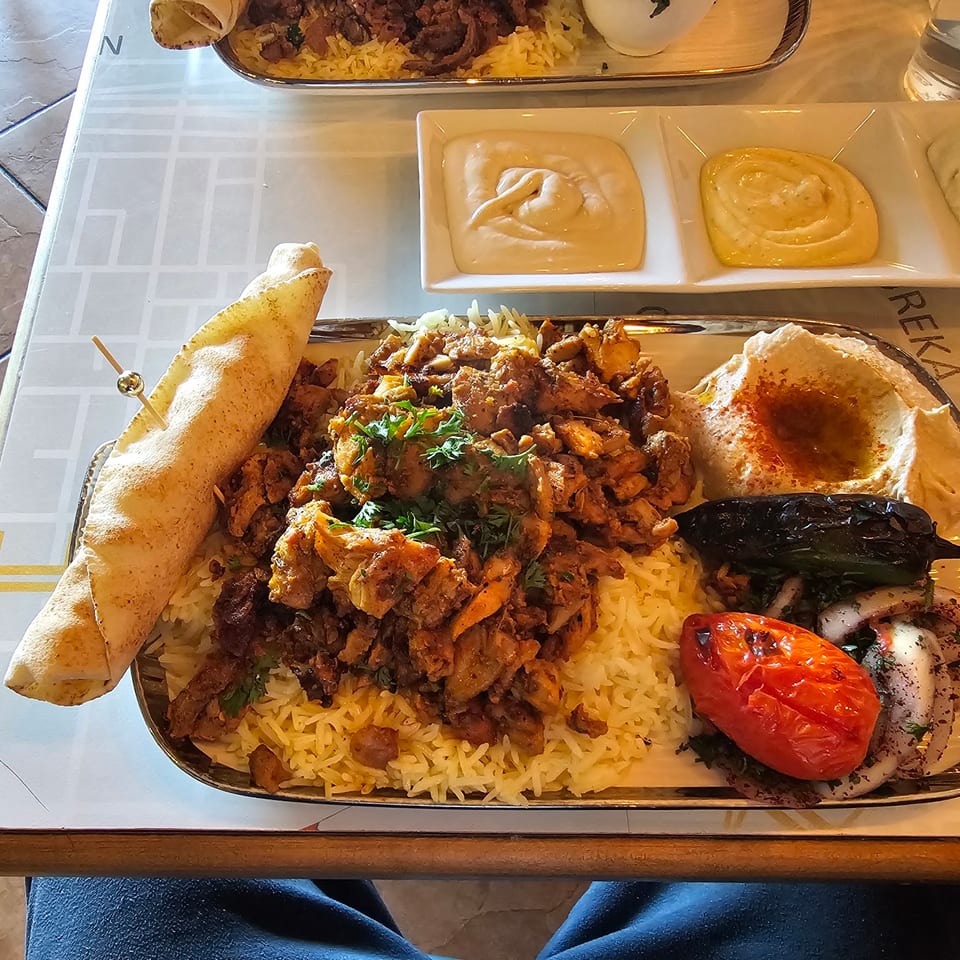 A metal platter of chicken shawarma, rice, pita, hummus, grilled vegetables, and sauces on a table.