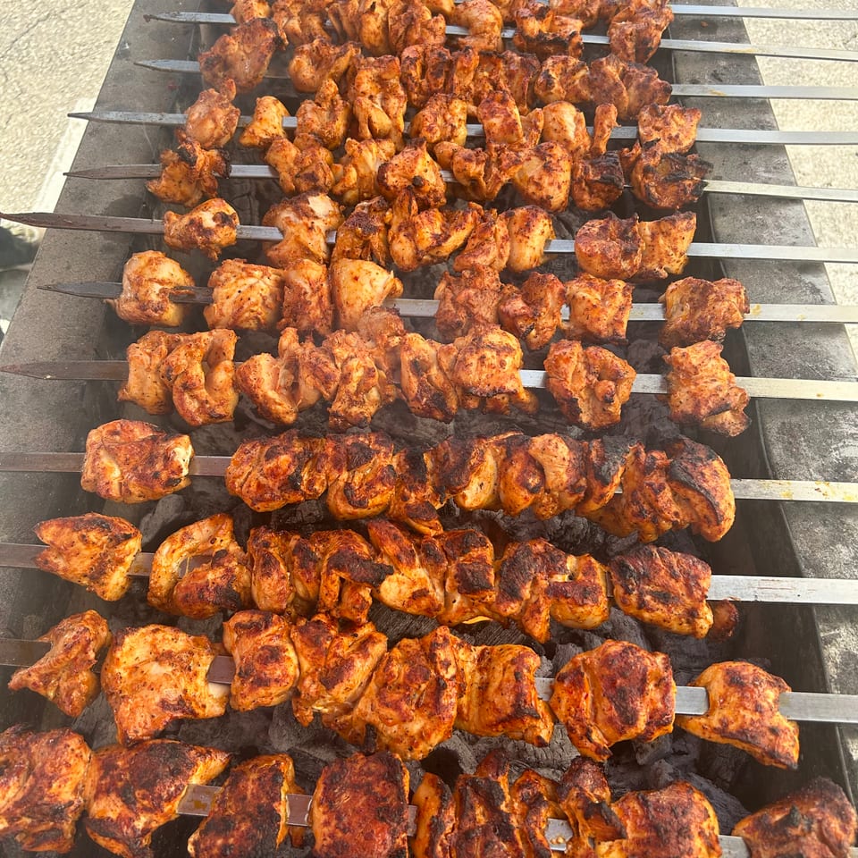 Chicken pieces on skewers grilling over hot coals, showing a large outdoor barbecue setup.