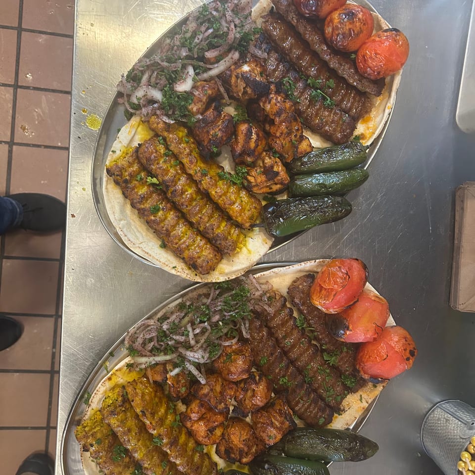 Two large platters of grilled meats, roasted tomatoes, peppers, and onions on flatbread.